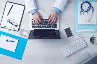doctors hands usng a laptop on a desk with other medical admin files