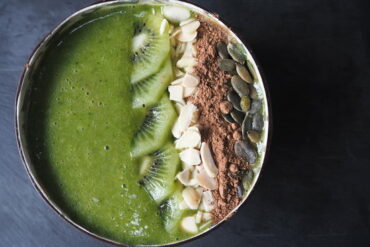 Close up picture of a green smoothie in a coconut bowl topped with slices of kiwi, flaked almonds, cacoa powder and pumpkin seeds