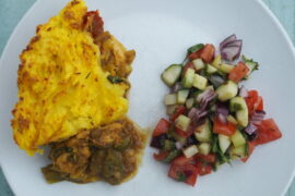White plate with yellow mashed potato on top of chunks of chicken in a jalfrezi sauce and a mixed dressed salad