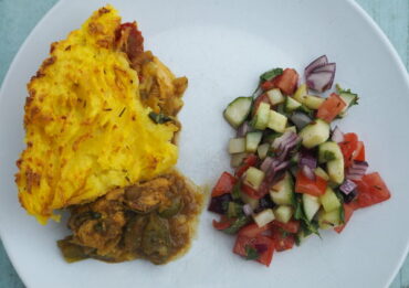 White plate with yellow mashed potato on top of chunks of chicken in a jalfrezi sauce and a mixed dressed salad