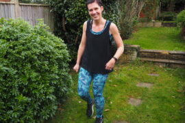 Smiling dark haired girl in graden next to a bush, wearing tropical blue leggings and black vest top
