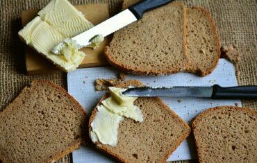 6 slices of brown bread on a chopping board wit two knives spreading lots of butter