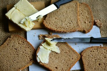 6 slices of brown bread on a chopping board wit two knives spreading lots of butter