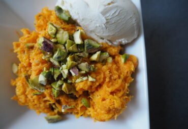 Close up of this carrot based dessert sprinkled with chopped pistachio nuts and a scoop of white vanilla ice cream in the top right hand corner