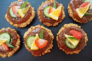 6 round flapjacks topped with Pimms strawberry jam and decorated with sliced strawberry, lemon, cucumber and mint leaf, on a dark grey slate background