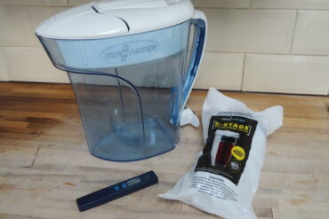 Water filter jug, filter cartidge in sealed plastic wrapping and a water meter test stick all sat on a wooden kitchen worktop with cream rectangular tiles on background