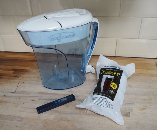 Water filter jug, filter cartidge in sealed plastic wrapping and a water meter test stick all sat on a wooden kitchen worktop with cream rectangular tiles on background
