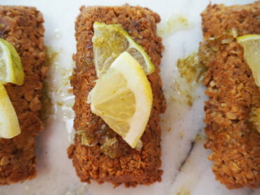 Close up of a rectangular flapjack drizzkled with gin and tonic flavoured syrup and topped with a quarter slice of lemon and lime. On a white marbled background