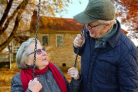 Older lady sat on a swing smiling up at an older man wearing a hat