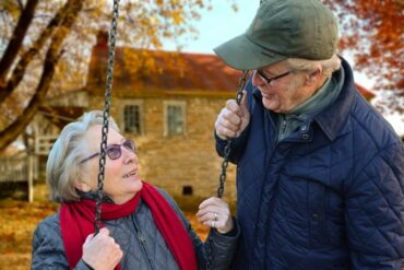 Older lady sat on a swing smiling up at an older man wearing a hat
