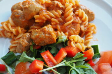 Close up of pasta twirls in tomato sauce with sausage meatballs and a side salad