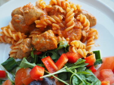 Close up of pasta twirls in tomato sauce with sausage meatballs and a side salad