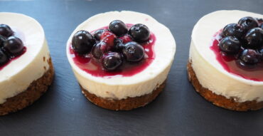 Close up of three smal round cheesecakes with a flapjack base and topped with blueberries and blueberry sauce on a dark slate background