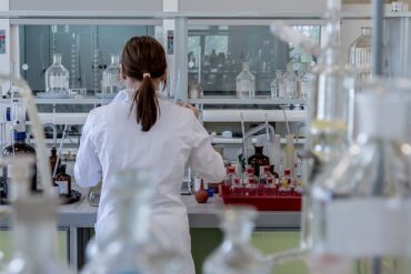 Back of female in white lab coat stood in a medical laboratory surrounded by medial equipment as she is studying the results from a clinical health trial