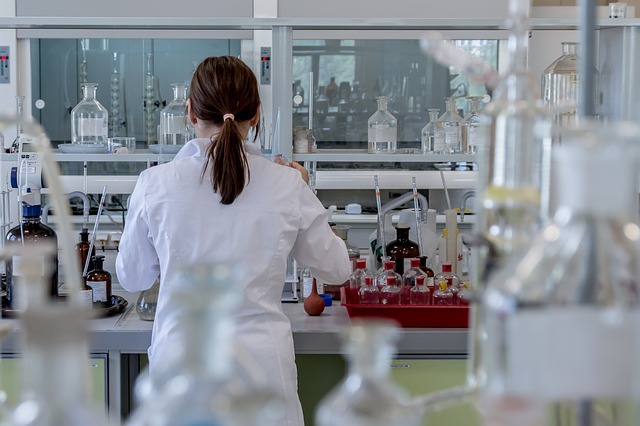 Back of female in white lab coat stood in a medical laboratory surrounded by medial equipment as she is studying the results from a clinical health trial