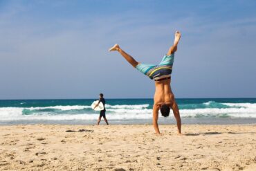 Man on a beach doinga handstand, man in the background holding a surfboard