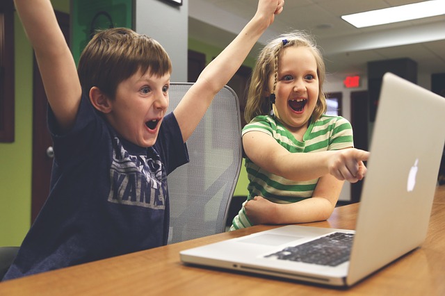 young boy and girl sat in front of an open laptop. Boy is cheering and girl is pointing at the screen and laughing. controversial subject matter perhaps.