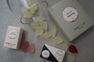Vinoos real wine gums scattered with packaging and wine glass. They are shaped like little bunches of grapes and come in red, rose and white wine flavours