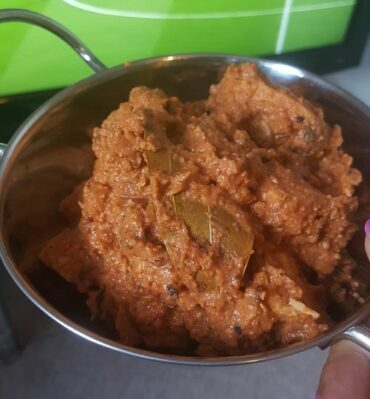 Close up of Makhani Murgh also known as Butter Chicken in a silver bowl in front of a close up of TV screen showing a football pitch
