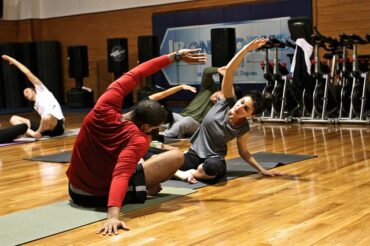 Couple sat facing each other in a gym, stretching to the side with their arms. Displays how exercising together can fomr intimate relationships