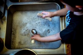 Close up of young boys arms, sleeves rolled up stood at a sink doing the washing up. Cutlery and sponge in hands with bubbly water in silver sink. Showing children should be using natural products.