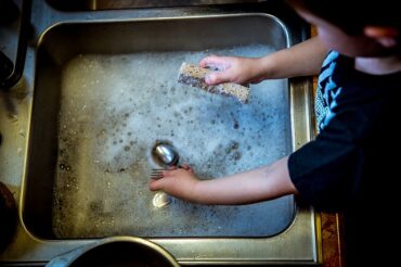 Close up of young boys arms, sleeves rolled up stood at a sink doing the washing up. Cutlery and sponge in hands with bubbly water in silver sink. Showing children should be using natural products.