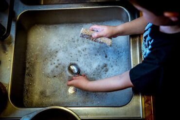 Close up of young boys arms, sleeves rolled up stood at a sink doing the washing up. Cutlery and sponge in hands with bubbly water in silver sink. Showing children should be using natural products.