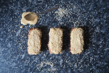 Three rectangular sesame seed flapjacks lined up in a row on a dark grey mottled kitchen worktop. Sesame seeds have been scattered around it and there is a teaspoon overspilling with tahini just above the flapjacks on the worktop.