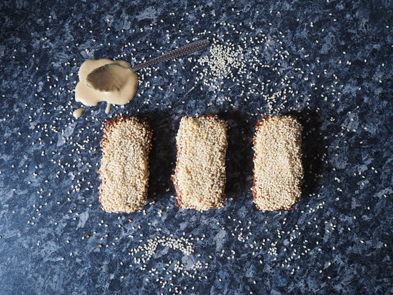 Three rectangular sesame seed flapjacks lined up in a row on a dark grey mottled kitchen worktop. Sesame seeds have been scattered around it and there is a teaspoon overspilling with tahini just above the flapjacks on the worktop.