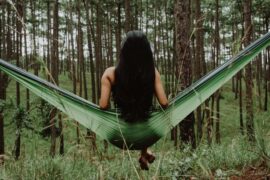 back of a long haired brunnet lady sat on a green hammock in the middle of a forest