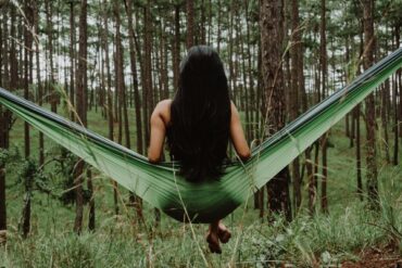 back of a long haired brunnet lady sat on a green hammock in the middle of a forest