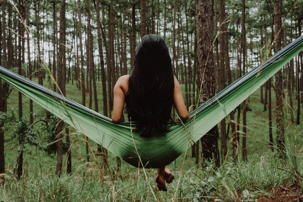 back of a long haired brunnet lady sat on a green hammock in the middle of a forest