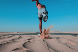 Topless man in shorts running across the sand. It's a blue skied day and sand is being flicked into the air as he runs freely to hep clear his mind