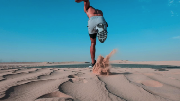 Topless man in shorts running across the sand. It's a blue skied day and sand is being flicked into the air as he runs freely to hep clear his mind