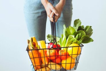 shopping basket filled with brightly coloured fruits and vegetables being held by a woman. You can only see her hand and jeans from waist down to knees