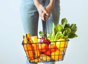 shopping basket filled with brightly coloured fruits and vegetables being held by a woman. You can only see her hand and jeans from waist down to knees