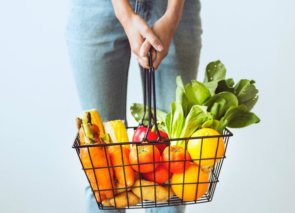 shopping basket filled with brightly coloured fruits and vegetables being held by a woman. You can only see her hand and jeans from waist down to knees