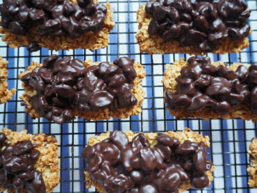 Close up of rectangular flapjack topped with dark chocolate raisins on a wire cooling rack with blue and white striped background