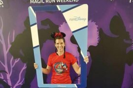 Blogger Becky Stafferton stood in front of Disneyland Paris purple run weekend back drop holding a blue selfie frame. She is wearing sparkly Minnie mouse ears and a red Disney half marathon run t-shirt and jeans. Her hair is in two plaits.