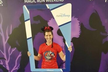 Blogger Becky Stafferton stood in front of Disneyland Paris purple run weekend back drop holding a blue selfie frame. She is wearing sparkly Minnie mouse ears and a red Disney half marathon run t-shirt and jeans. Her hair is in two plaits.