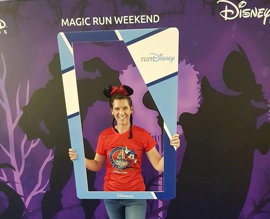 Blogger Becky Stafferton stood in front of Disneyland Paris purple run weekend back drop holding a blue selfie frame. She is wearing sparkly Minnie mouse ears and a red Disney half marathon run t-shirt and jeans. Her hair is in two plaits.