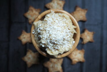Small wooden bowl of sweet coconut hummus topped with sprinkled coconut. In the dark background are some star chaped cardamom coconut biscuits going around the bowl