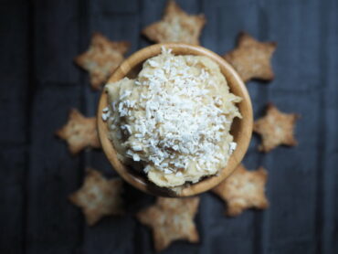 Small wooden bowl of sweet coconut hummus topped with sprinkled coconut. In the dark background are some star chaped cardamom coconut biscuits going around the bowl