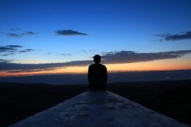 man sitting at the end of a pier. It is dusk and the sun has recently set, the sky is dark blue fading to orange down to the dark water in front of him. He is contemplating his life and thiking about quitting a bad habit.