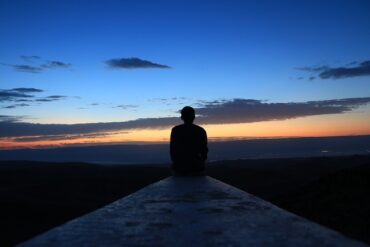 man sitting at the end of a pier. It is dusk and the sun has recently set, the sky is dark blue fading to orange down to the dark water in front of him. He is contemplating his life and thiking about quitting a bad habit.