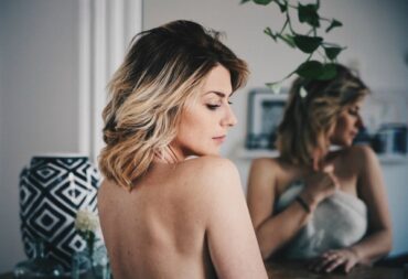 Woman in her thirties sat in front of a mirror with her back to the camera. She is topless and looking down to the floor feeling confident and beautiful in her own skin