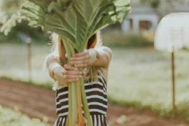 Blonde haired lady in a black and white striped dress holding out a bing bunch of rhubarb so that it covers her face. She looks as if she is stood in an allotment and is showing people how you can take better care of your health