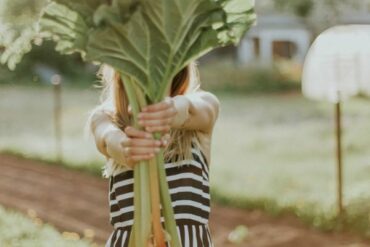 Blonde haired lady in a black and white striped dress holding out a bing bunch of rhubarb so that it covers her face. She looks as if she is stood in an allotment and is showing people how you can take better care of your health