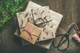 Threee different sized square presents stacked on top of one another. Eacgh one is wrapped in brown packing paper and looks very natural. There is some pine and a pair of scissors next to them and they are on a wodden floor.