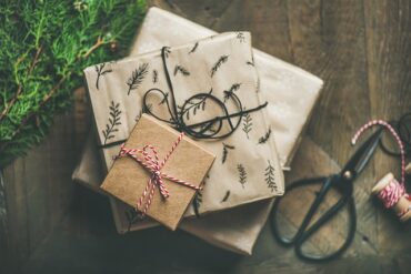 Threee different sized square presents stacked on top of one another. Eacgh one is wrapped in brown packing paper and looks very natural. There is some pine and a pair of scissors next to them and they are on a wodden floor.